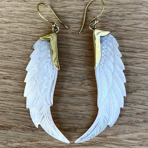 May include: A pair of white wing-shaped earrings with intricate feather detailing. The earrings have gold-colored metal accents and ear wires. The wings are carved with delicate lines, creating a textured appearance. The earrings are displayed on a wooden surface.