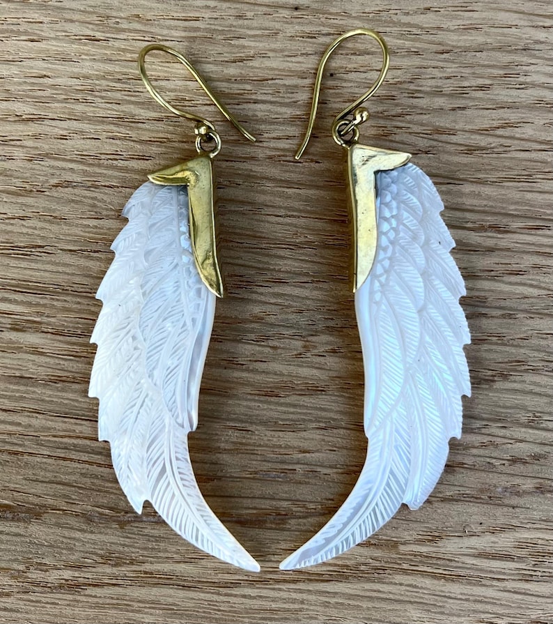May include: A pair of white wing-shaped earrings with intricate feather detailing. The earrings have gold-colored metal accents and ear wires. The wings are carved with delicate lines, creating a textured appearance. The earrings are displayed on a wooden surface.