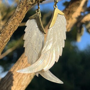 May include: A pair of earrings featuring iridescent, carved white wing designs with gold-colored metal accents. The earrings are suspended from gold-colored hooks, and are displayed against a natural, sunlit background.