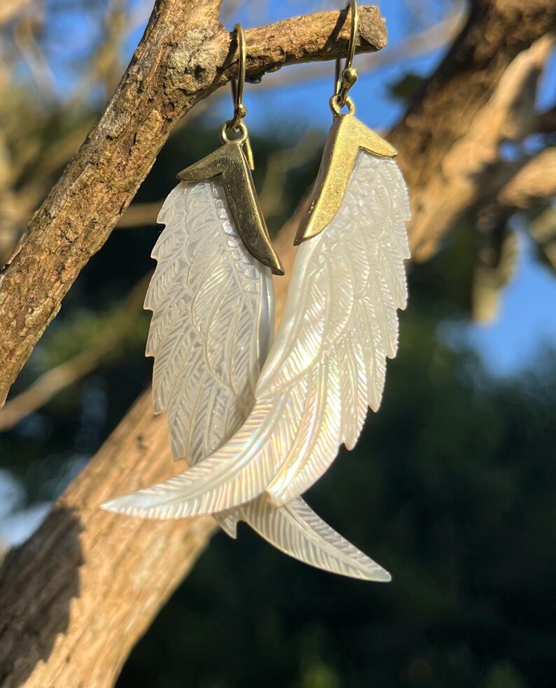 May include: A pair of earrings featuring iridescent, carved white wing designs with gold-colored metal accents. The earrings are suspended from gold-colored hooks, and are displayed against a natural, sunlit background.