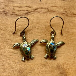 May include: A pair of gold-toned metal earrings shaped like sea turtles. Each turtle features an inlaid shell section made of iridescent abalone shell with blue and green hues. The earrings hang from simple curved gold-toned metal ear wires.
