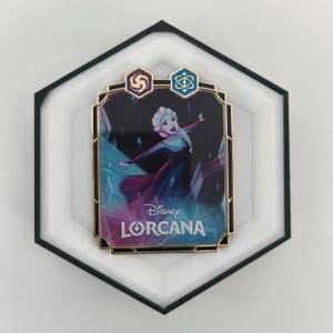 Könnte beinhalten: Ein Disney Lorcana Pin mit Elsa aus Frozen. Der Pin hat einen goldenen Rand und ein rechteckiges Design mit dem Disney Lorcana Logo unten. Der Hintergrund ist eine Mischung aus Blau, Lila und Weiß.