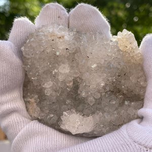 May include: A cluster of clear quartz crystals, held in white gloves, with a sparkling, crystalline structure. The quartz formation has a light gray hue with some areas of white and is set against a blurred green background.