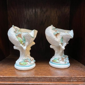 May include: Two white ceramic seashell-shaped bowls. Each bowl is supported by a tree-like base with blue and green accents. The bowls have a textured surface and a light blue trim.