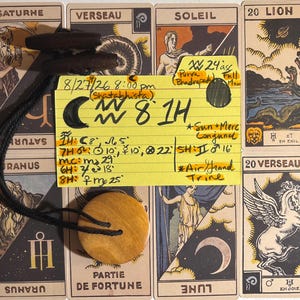 May include: A yellow index card with handwritten astrological notes and symbols, including the text 8/27/26 8:00 pm, placed over several tarot cards. A round, light-colored wooden pendant with a black cord is attached to the card, along with two dark toggles.