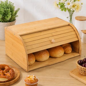 May include: A rectangular bamboo bread bin with a roll-top lid featuring a small wooden knob. The light brown wooden container has a curved side profile and is open at the front, revealing three golden-brown baked bread rolls inside the storage unit.