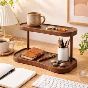 May include: A two-tiered wooden desk organiser with rounded edges, featuring a top shelf holding a ceramic mug and a magnifying glass, and a bottom shelf containing a brown leather notebook, a pen, a speckled white pen holder, and three stones.