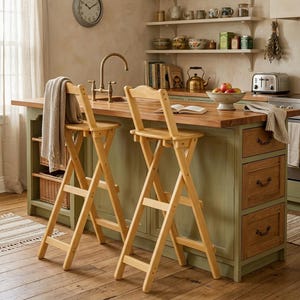 May include: Two light-toned wooden folding bar stools with X-shaped legs and backrests, positioned in front of a kitchen island with a wooden countertop, sage green cabinetry, and three drawers with dark metal pulls.