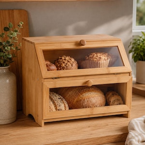 May include: A two-tier bamboo bread box with clear glass windows and small wooden knobs. The top compartment holds muffins and small rolls, while the bottom compartment contains a large round loaf of bread and sliced bread pieces.