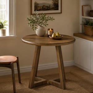 May include: A round wooden table with a smooth top and three angled legs that meet at the base. On the table sits a white ribbed ceramic vase containing green leafy branches, a lit candle in a glass jar, and a small bowl holding two yellow pears.