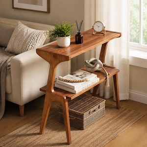 May include: A wooden console table with a smooth, warm-toned finish, featuring a rectangular top and a lower shelf. The table has angled legs and supports. On the top surface sits a small potted green plant, a dark glass diffuser, and a clock.