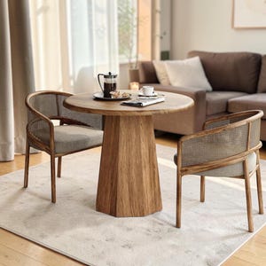 May include: A round wooden dining table with a faceted pedestal base paired with two chairs featuring curved wooden frames, woven cane backrests, and light grey upholstered seat cushions. The table surface holds a glass carafe and a white cup.
