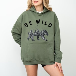 May include: An olive green hooded sweatshirt with the words "BE WILD" in black block letters across the top. Below the text is a black and white graphic of a bear with a forest scene inside its silhouette.