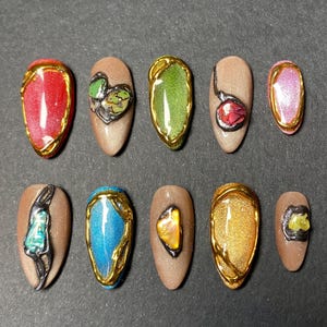 May include: A set of ten almond-shaped press-on nails featuring various designs. Some nails have solid metallic or glittery finishes in red, green, blue, and gold, while others are beige with raised, textured accents and metallic gold or black trim.