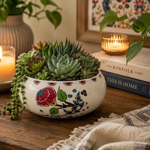 May include: A round white ceramic planter filled with a variety of green succulents, including a trailing string of pearls. The planter features a hand-painted floral pattern with red, blue, and green colors around the exterior rim and body.
