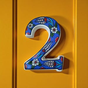 May include: Decorative ceramic number 2 in blue, white, and red floral design. The number is mounted on a yellow surface. The floral pattern includes white flowers, red accents, and green leaves. The number is a decorative item.