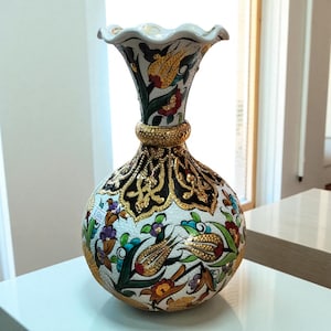 Può includere: Un vaso decorativo in ceramica con base bianca, caratterizzato da intricati motivi floreali in rosso, giallo, verde e blu. Il vaso ha un bordo increspato e una fascia dorata attorno al collo. Il design include dettagli dorati e filigrana nera.