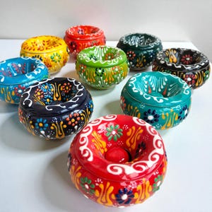 May include: A selection of vibrant ceramic ashtrays. Each ashtray is round with a lid and adorned with detailed floral designs in shades of red, yellow, blue, green, and black. The ashtrays are displayed on a white surface.
