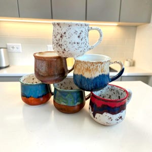 Op de afbeelding: Een stapel van zes keramische mokken met gevarieerde afwerkingen. Eén is wit met bruine spikkels, een andere is effen bruin, één heeft een blauw en beige druipglazuur, één is blauw en oranje, één groen en bruin, en één is wit met rode en blauwe druppels.