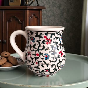 May include: A white ceramic mug with a floral pattern in red, blue, and green. The mug has a curved handle and a black rim. A small plate of cookies sits nearby on a light green tray.