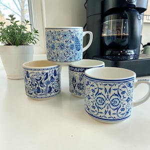 Puede incluir: Cuatro tazas de cerámica con diseños florales y de pájaros en azul y blanco. Una taza tiene asa, mientras que las otras son cilíndricas. Las tazas están dispuestas sobre una superficie blanca cerca de una cafetera y una planta en maceta.
