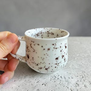以下が含まれることがあります： 丸みを帯びたボディとハンドルが付いた、小さな白いセラミックマグ。マグには濃い茶色の斑点があります。マグは手で持たれており、背景はニュートラルグレーです。
