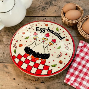 Może przedstawiać: Okrągła ceramiczna pokrywka z kremowym tłem i czerwono-białym wzorem w kratkę u dołu. Przedstawia trzy stylizowane kurczaki oraz napis Egg-hausted czcionką w kolorze czarnym, otoczony małymi czerwonymi kwiatami i zielonymi liśćmi.