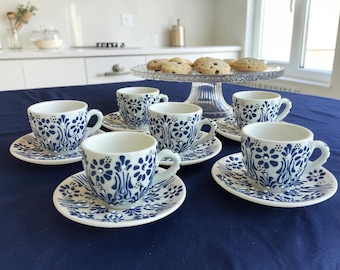 May include: A set of four white ceramic espresso cups and matching saucers, each decorated with a blue floral pattern and small raised white dots. The cups are arranged on a dark blue tablecloth, with a glass cake stand holding cookies behind.