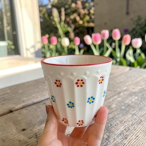 Op de afbeelding: Een witte keramieken beker met een geribbelde, gecanneleerde buitenkant en een rode rand. Het oppervlak is versierd met een herhalend patroon van kleine, handgeschilderde bloemen met rode en blauwe blaadjes en gele harten.