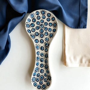 May include: A white ceramic spoon rest with a blue and navy eye pattern. The spoon rest is oval-shaped with a long handle. A blue cloth and a beige cloth are in the background.