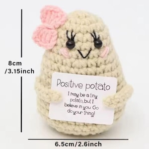 Puede incluir: Una muñeca de patata tejida a ganchillo, de aproximadamente 8 cm de alto y 6,5 cm de ancho, con un lazo rosa y una cara sonriente. Sostiene un letrero que dice "Positive potato".