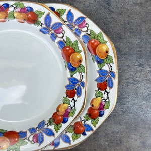May include: Two white ceramic plates with a gold rim and a blue and white floral design featuring fruit. The plates have a white center and are decorated with a pattern of red, yellow, and blue fruit.