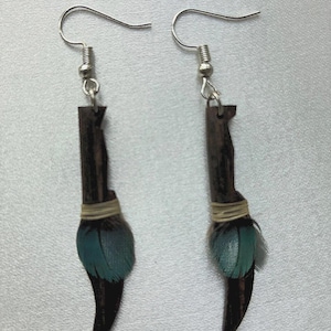 Pode incluir: Um par de brincos artesanais. Cada brinco apresenta um elemento de madeira castanha escura, um aglomerado de penas verde-azuis e uma parte inferior curva castanha escura. Os brincos penduram de ganchos prateados.