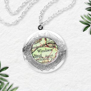 Puede incluir: Un collar con colgante de medallón redondo plateado. El medallón presenta un diseño floral grabado en los bordes y un mapa de Windsor, Inglaterra, en el interior. La cadena del collar también es plateada.