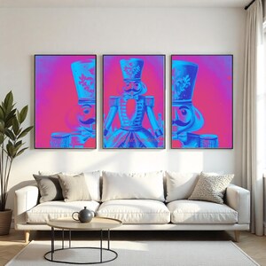 Peut inclure: Trois impressions encadrées représentant un soldat casse-noisette bleu et rose dans un style pop art. Les casse-noisettes sont représentés de manière détaillée et stylisée, avec un accent sur leurs uniformes et leurs chapeaux. L'œuvre est exposée au-dessus d'un canapé blanc.