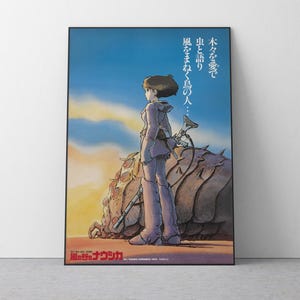 Puede incluir: Póster enmarcado de una película de animación, mostrando un personaje en un paisaje desértico. El personaje, con un atuendo lila claro, está de espaldas. Un gran insecto y texto japonés son visibles en el fondo.