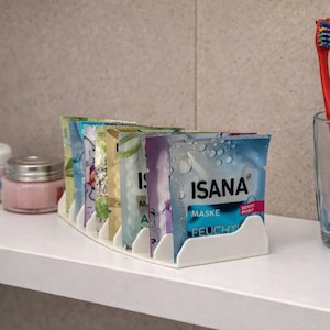 Könnte beinhalten: Ein weißes Regal mit Hautpflegeprodukten. Ein weißer Organizer enthält ISANA Gesichtsmasken. Ein Glas enthält drei Zahnbürsten mit roten, blauen und rosa Griffen. Weitere Artikel sind eine Creme und ein kleines rosa Glas.