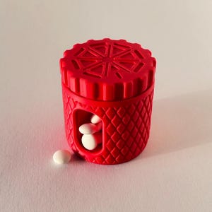 Könnte beinhalten: Ein leuchtend roter Pillenbehälter mit einem Diamantmuster und einem geometrischen Design auf dem Deckel. Der Behälter enthält mehrere weiße Pillen, wobei einige Pillen außerhalb sichtbar sind.