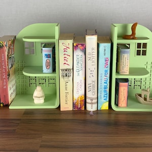 Peut inclure: Une collection de livres et d'objets décoratifs disposés sur une surface en bois. Les livres sont de tailles et de couleurs variées, avec des titres visibles. Deux serre-livres en forme de maison vert clair soutiennent certains livres. Petits objets décoratifs.