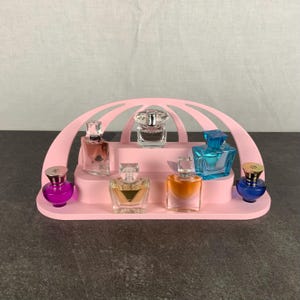 Könnte beinhalten: Ein rosa Präsentationsständer mit mehreren Miniatur-Parfümflaschen. Die Flaschen haben verschiedene Formen und Farben, darunter Lila, Blau, Orange und Klar. Der Ständer hat eine gewölbte Oberseite und ein mehrstufiges Design.