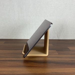 Könnte beinhalten: Ein schwarzes Tablet steht auf einem beigefarbenen Ständer. Der Ständer ist abgewinkelt und bietet einen Betrachtungswinkel für das Tablet. Der Ständer besteht aus einem festen Material. Der Hintergrund ist ein hellbeiger Stoff und eine dunkelbraune Holzoberfläche.