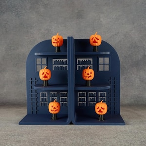 Può includere: Un espositore a forma di arco blu scuro con due sezioni verticali e diversi ripiani. Sei piccole figure di zucca arancioni con espressioni intagliate sono posizionate sui ripiani, ognuna montata su una base cilindrica nera.