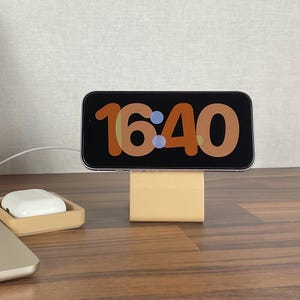 Könnte beinhalten: Ein Smartphone mit der Uhrzeit 16:40 liegt auf einem beigen Ladeständer. Das Telefon hat einen schwarzen Bildschirm und einen silbernen Rahmen. Ein weißes kabelloses Kopfhörer-Etui befindet sich in einer passenden beigen Schale, mit einem Laptop im Vordergrund.