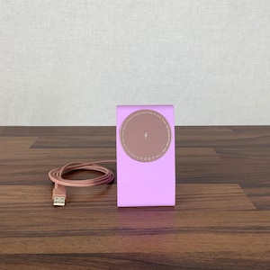 Könnte beinhalten: Ein pinkfarbenes kabelloses Ladegerät mit einem braunen Lade-Pad und einem Blitzsymbol. Ein passendes pinkfarbenes USB-Kabel ist neben dem Ladegerät aufgerollt. Das Ladegerät steht auf einer Holzoberfläche.