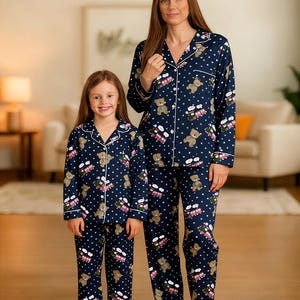 Könnte beinhalten: Passende marineblaue Pyjama-Sets mit Teddybär- und Zugmuster. Die Pyjamas haben weiße Tupfen und weiße Paspeln und werden von einer Frau und einem Kind getragen. Das langärmlige Oberteil und die Hose sind aus weichem Material.
