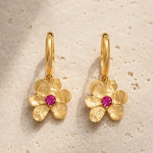 May include: A pair of gold hoop earrings featuring a dangling five-petal flower charm with a textured finish and a small, round, vibrant pink gemstone set in the center of each flower. The earrings have a polished gold tone and a hinged closure.