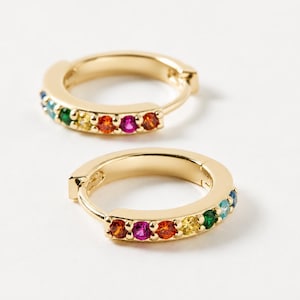 Peut inclure: Une paire de créoles dorées ornées d'une rangée de petites pierres précieuses multicolores serties le long de la courbe avant. Les pierres présentent un spectre de couleurs vives incluant le rouge, l'orange, le jaune, le vert et le bleu.