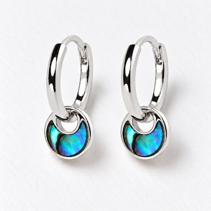 May include: A pair of silver hoop earrings featuring a dangling circular charm. Each charm contains a crescent-shaped inlay of iridescent abalone shell displaying vibrant blue and green tones set within a polished silver frame.