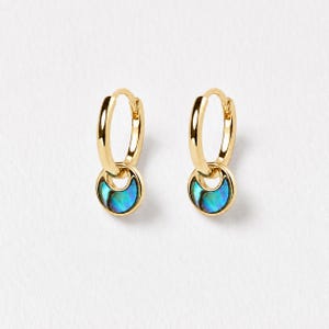May include: A pair of gold hoop earrings featuring a small, circular pendant charm. Each charm contains a polished, iridescent abalone shell inlay that displays vibrant shades of blue, green, and purple, set within a smooth, polished gold frame.