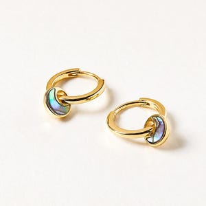 May include: A pair of gold-coloured hoop earrings, each with a small, iridescent, half-moon charm. The charms have blue, green, and purple tones. The earrings are displayed on a white background.
